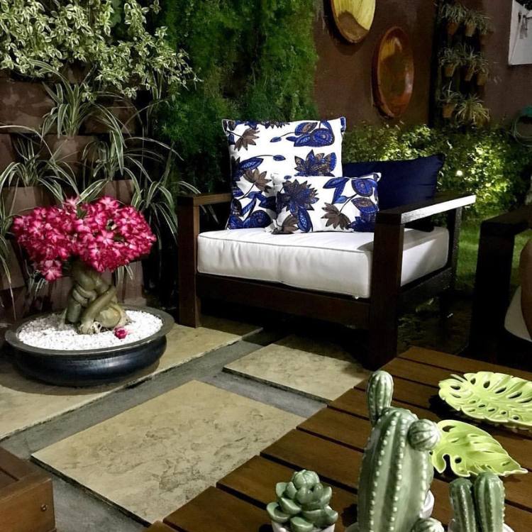 Varanda com rosa do deserto em vaso preto, outras plantas, poltrona marrom de estofado branco e decorações verdes