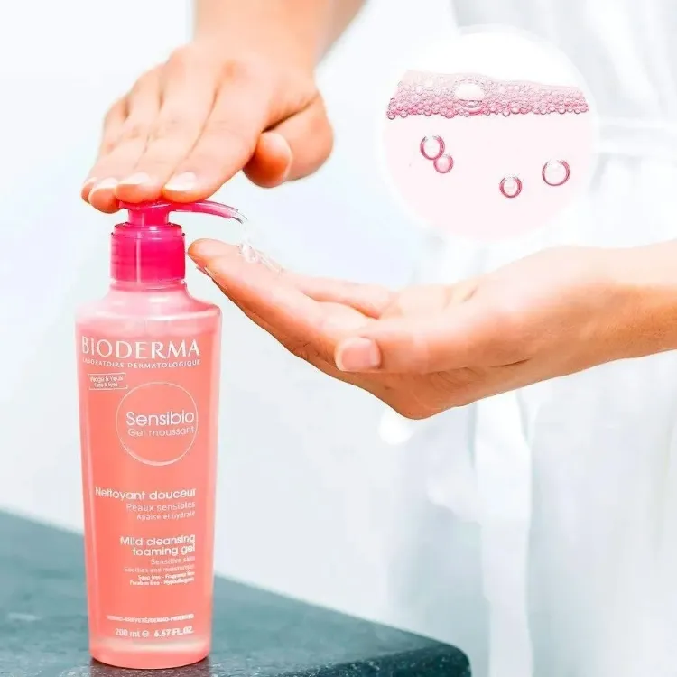 A imagem mostra um sabonete para pele sensível, o Bioderma Sensibio Gel Moussant, sendo dispensado de um frasco rosa para a mão. Bolhas de sabão rosadas flutuam em destaque no canto superior direito, ilustrando a textura espumosa do produto de limpeza suave.







