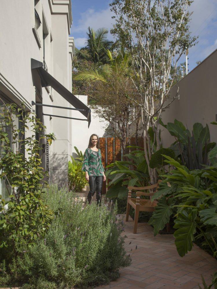 Imagem de uma área externa de uma casa, com diversas plantas e uma mulher de pele clara ao fundo, usando calça preta e blusa verde.