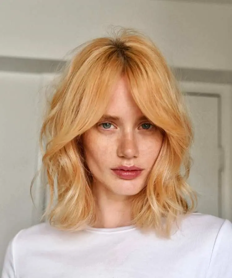 Mulher com cabelo médio loiro acobreado, corte em camadas suaves com franja cortina dividida ao meio. Ela veste uma blusa branca e olha diretamente para a câmera. O ambiente interno possui fundo neutro, destacando o penteado elegante.