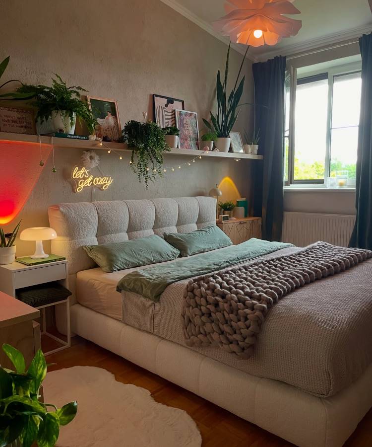 Uma cama queen size com uma cabeceira estofada e roupa de cama verde e cinza. Prateleiras acima exibem plantas, quadros e luzes de neon. O quarto aconchegante possui iluminação suave e plantas em vasos, criando um ambiente tranquilo.