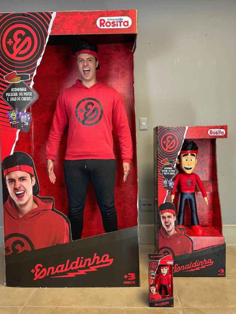 Enaldinho usando moletom vermelho de sua amrca e calça preta dentro de embalagem gigante de seu boneco e versões de seu boneco ao lado