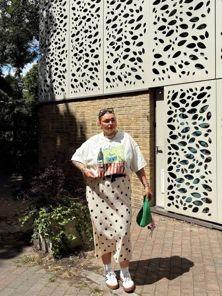 Uma mulher sorridente posa em frente a um prédio com fachada de tijolos e painéis vazados. Ela veste uma camiseta branca com estampa de mesa, uma saia midi com estampa de bolinhas e tênis brancos. Ela segura uma bolsa verde.