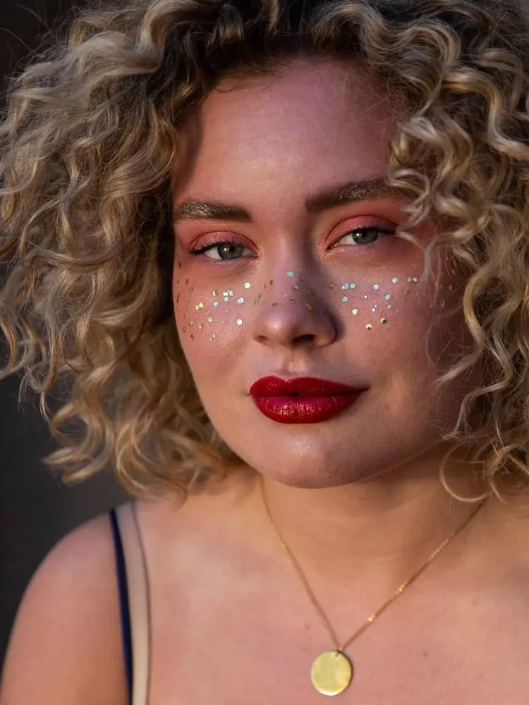 Mulher de pele clara com batom vermelho, glitter dourado na sobrancelha e strass no rosto 
