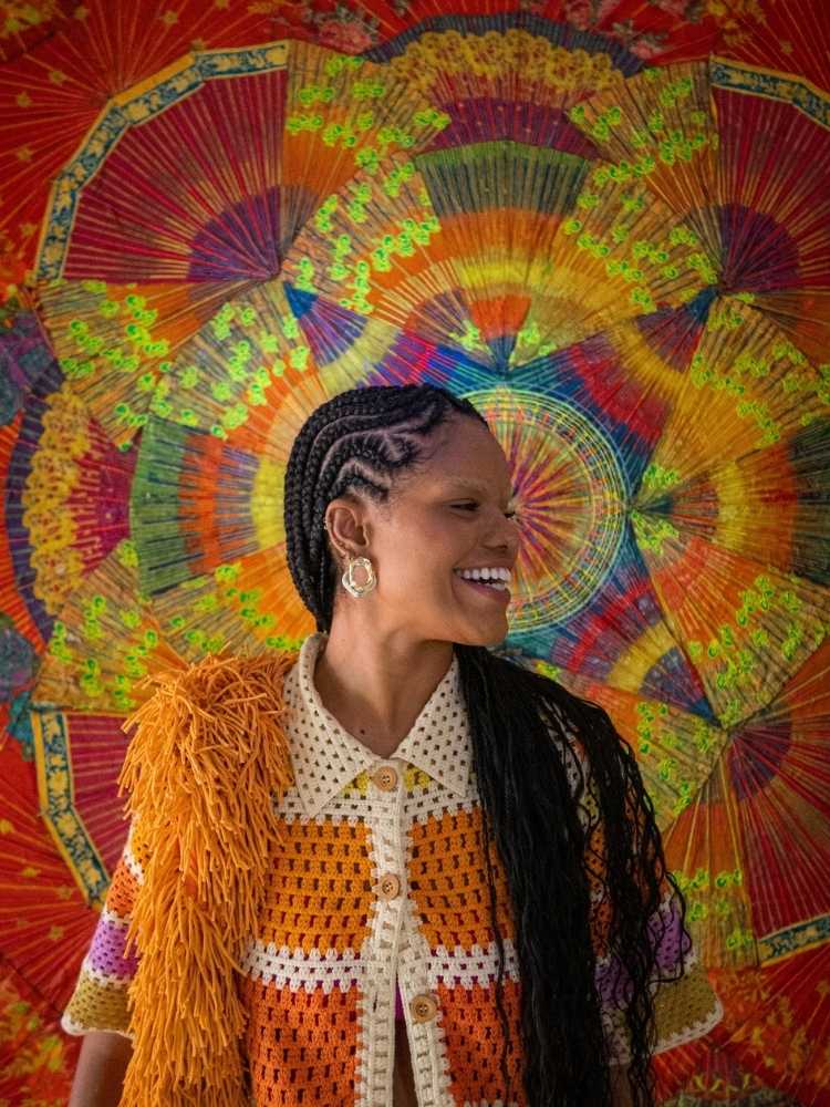 Jucy Ramos de roupa de crochê branca e laranja, em fundo colorido, usando trança nagô