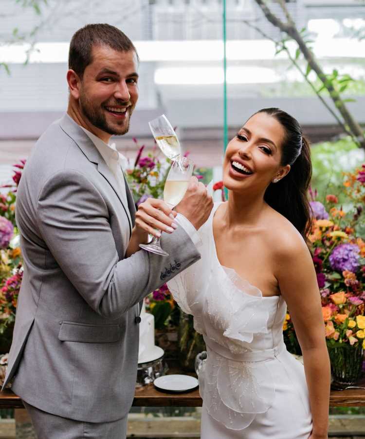 Casal Gustavo Marsengo e Laís Caldas sorridente brinda com taças de champanhe em cerimônia luxuosa. Noiva usa vestido branco com detalhes delicados, enquanto noivo veste terno cinza claro. Flores coloridas decoram o ambiente, destacando a elegância dos casamentos de famosos 2025 em cenário encantador.
