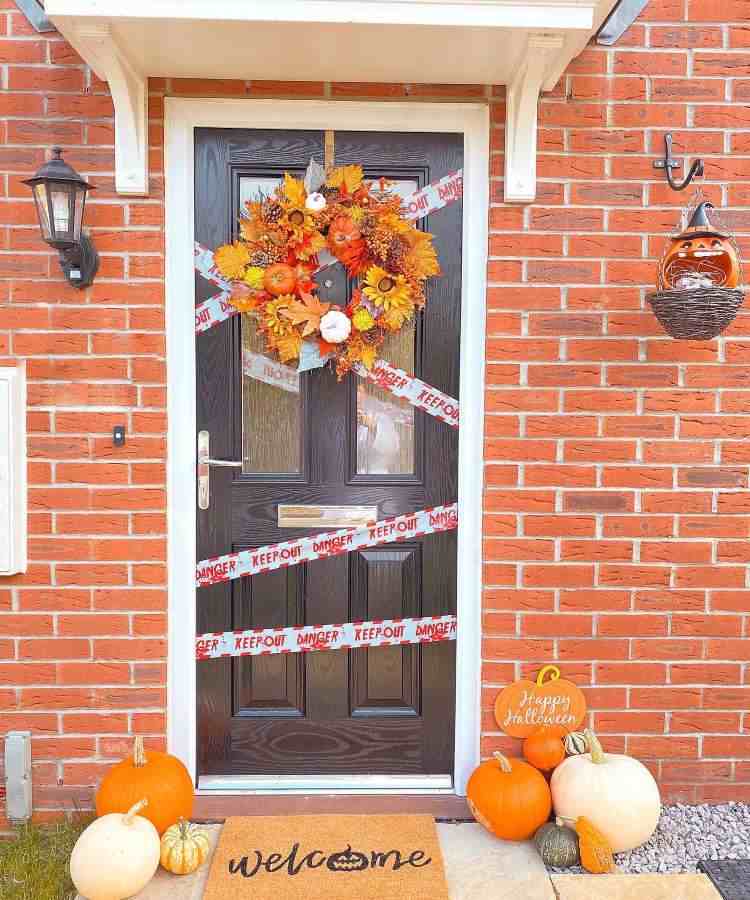Entrada de casa com tijolos à vista, porta de madeira com fita, guirlanda com flores laranjas, tapete de Halloween e abóboras