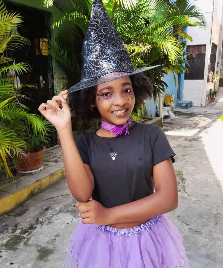 Garota fantasiada para o Halloween: Cores predominantes: preto e lilás. Usa: cropped, saia de tule, gargantilha de tecido, chapéu pontudo