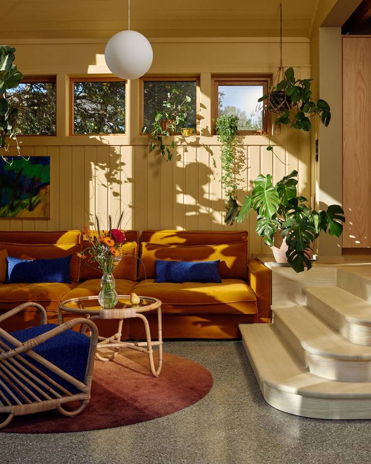A imagem mostra uma sala de estar aconchegante e bem iluminada. Um sofá amarelo com almofadas azuis está posicionado ao lado de uma escada em espiral branca. Uma mesa de centro redonda com um vaso de flores coloridas está na frente do sofá. Há plantas penduradas perto das janelas que permitem a entrada abundante de luz solar. O chão é coberto por um tapete redondo e escuro, contrastando com o piso claro. Uma pintura abstrata colorida adorna a parede à esquerda da imagem. A sala é iluminada pelo sol, com um sofá amarelo, plantas penduradas, uma mesa de centro e uma escada em espiral.