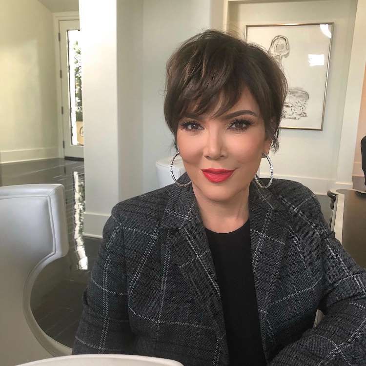 Kris Jenner sorri com um batom vermelho vibrante e grandes brincos de argola. Ela veste um blazer xadrez cinza e seu cabelo é um corte pixie castanho, em um ambiente bem iluminado.