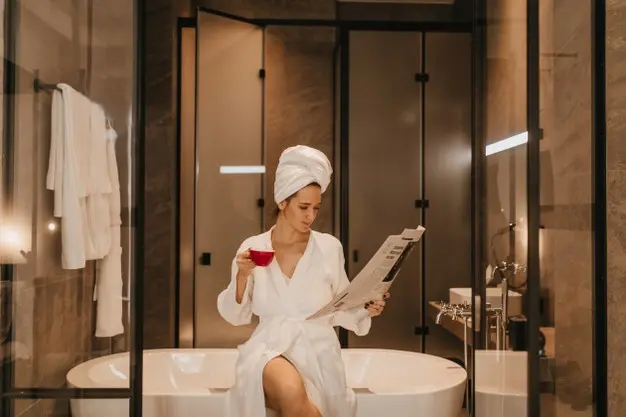 Mulher de pele clara em banheiro marrom luxuoso, sentada à beira de banheira, usando toalha na cabeça, roupão branco e lendo jornal enquanto segura xícara vermelha  