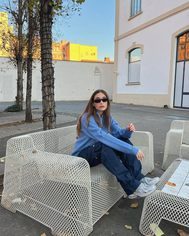 Jovem garota usando calça cargo, camisa de frio azul e tênis, sentado em sofá de ferro em pátio de casa.