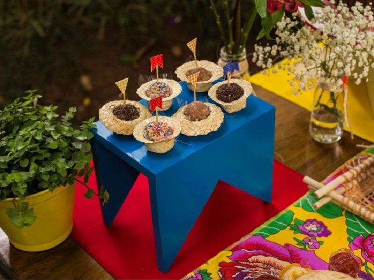 A imagem representa mini chapéus de palha com docinhos dentro, como brigadeiro e beijinho, em mesa decorada de festa junina
