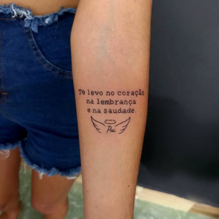 A imagem mostra uma tatuagem no antebraço de uma pessoa com a frase "Te levo no coração na lembrança e na saudade.", seguida pela palavra "Pai" e um par de asas de anjo com uma auréola. A tatuagem é em preto e a pele parece clara. A pessoa veste shorts jeans.







