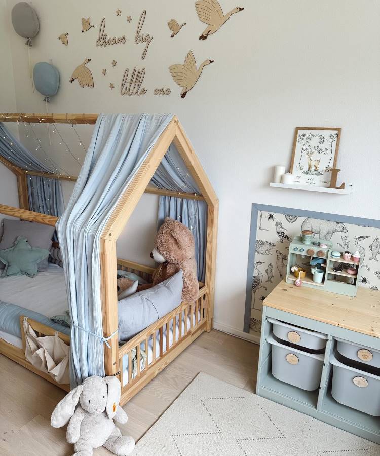 O quarto infantil apresenta uma cama em formato de casinha de madeira com cortinas azul-claro e luzes decorativas. Um grande urso de pelúcia está sobre a cama. Na parede, adesivos de gansos voando e a frase “dream big little one”. Uma estante organiza brinquedos e utensílios.