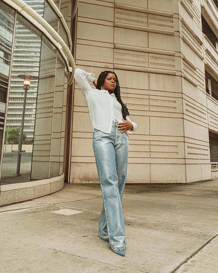 Mulher negra usando camisa branca, calça jeans metalizada, com sling back, em local público com construção, possivelmente de um prédio.
