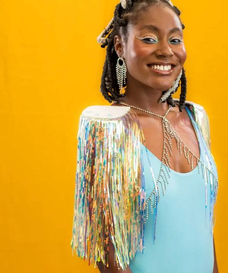 Uma mulher sorridente usa um body azul com ombreiras decoradas com franjas brilhantes e coloridas. Ela exibe maquiagem vibrante, brincos longos e cabelo estilizado com tranças. O fundo amarelo destaca a atmosfera alegre e carnavalesca da imagem.