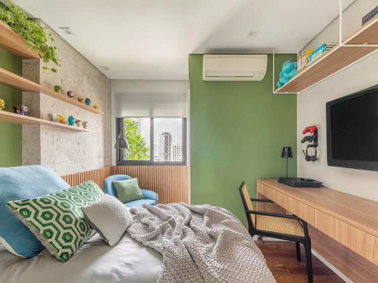 Quarto do filho mais novo, cores verde e tons neutros, cama com enxoval verde, azul e cinza. Poltrona azul com almofada verde. A cama tem na frente uma parede com televisão, vídeo game, cadeira com mesa e acima prateleira com objetos. Acima da cabeceira da cama, prateleiras com plantas e bichinhos decorando