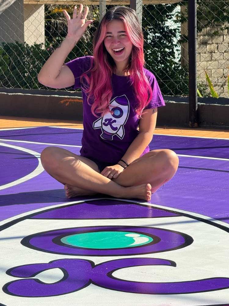 Katlen Martins vestindo shorts e camiseta roxa com foguete sentada no chão pintado de roxo com foguete