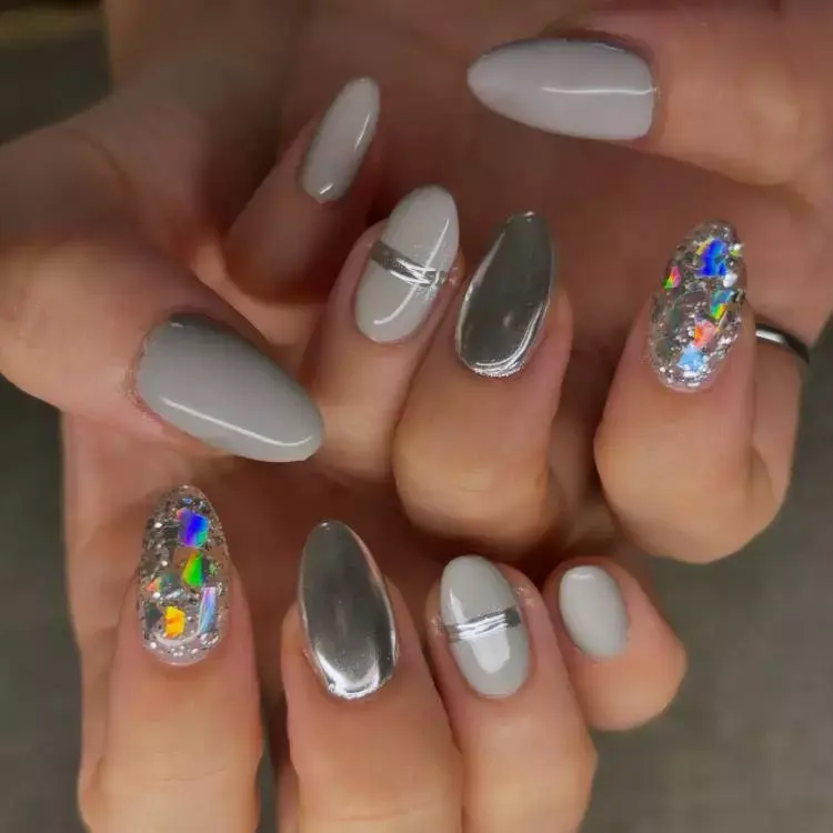 A imagem mostra unhas longas e ovais com um design sofisticado. As unhas são pintadas em tons de cinza e prateado, com algumas apresentando um acabamento metálico espelhado. Duas unhas possuem decoração com glitter holográfico, criando um efeito brilhante e colorido. O design inclui linhas prateadas horizontais em algumas unhas, adicionando um toque moderno e elegante.


