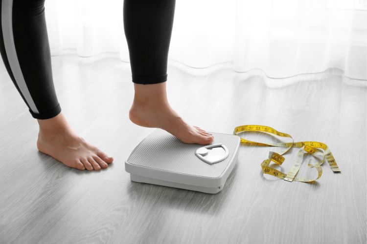 Pessoa de pele clara usando calça legging preta, pisando levemente em balança, ao lado de fita métrica amarela no chão
