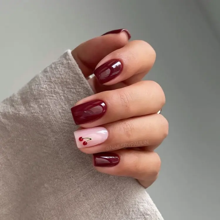 Mão de pele clara com unhas quadradas em vinho e anelar com rosa claro e desenho de cereja 