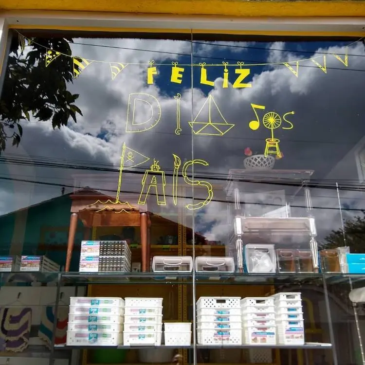 A imagem mostra uma vitrine de loja com a mensagem "FELIZ DIA DOS PAIS" escrita em adesivo amarelo no vidro. Acima, há uma guirlanda de bandeirinhas amarelas. Prateleiras com diversos produtos brancos organizados são visíveis, e o céu nublado se reflete no vidro.