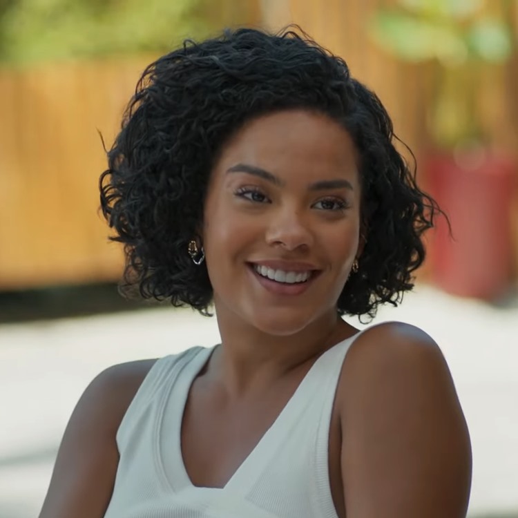 Bella Campos como Maria de Fátima em Vale Tudo. Mulher sorrindo com cortes de cabelo de Vale Tudo, cabelo cacheado curto, blusa branca, brincos dourados, em cenário externo iluminado com fundo de madeira.





