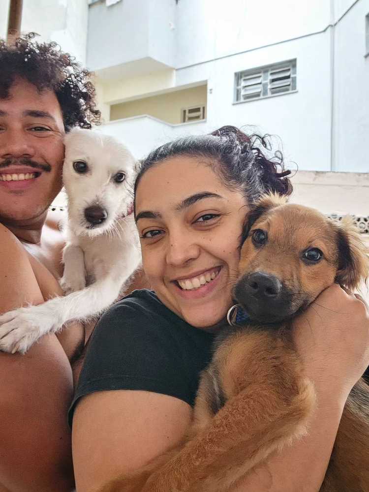 Lucas Henrique com vira-lata branco no colo e Camila, uma mulher de pele clara e cabelo escuro, abraçada com um vira-lata caramelo. 