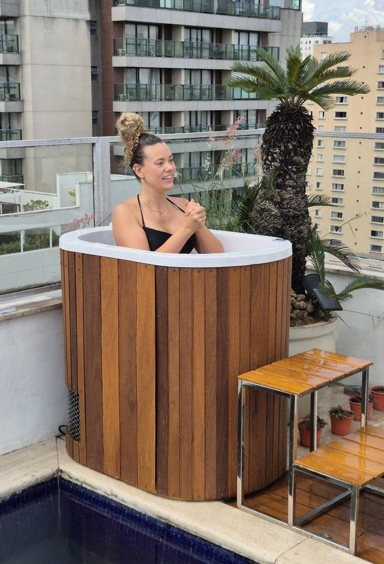 Mulher sorridente relaxando em uma banheira de ofurô de madeira em uma cobertura. A vista ao fundo mostra prédios e uma piscina. Ela está usando um biquíni preto e tem o cabelo preso.







