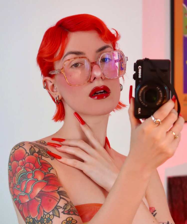Mulher de cabelo curto vermelho vibrante com ondas estilizadas, usando óculos de armação grande transparente. Maquiagem com batom vermelho brilhante, unhas longas vermelhas, e tatuagens florais coloridas no braço. Segura câmera DSLR e veste top laranja em ambiente iluminado.