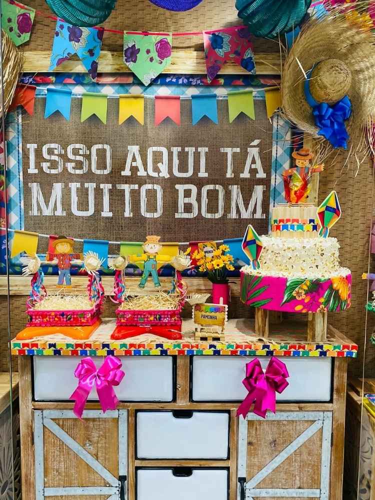 Vitrine com painel escrito "isso aqui tá muito bom", com bandeirinhas, palha e bolo fake de pipoca 