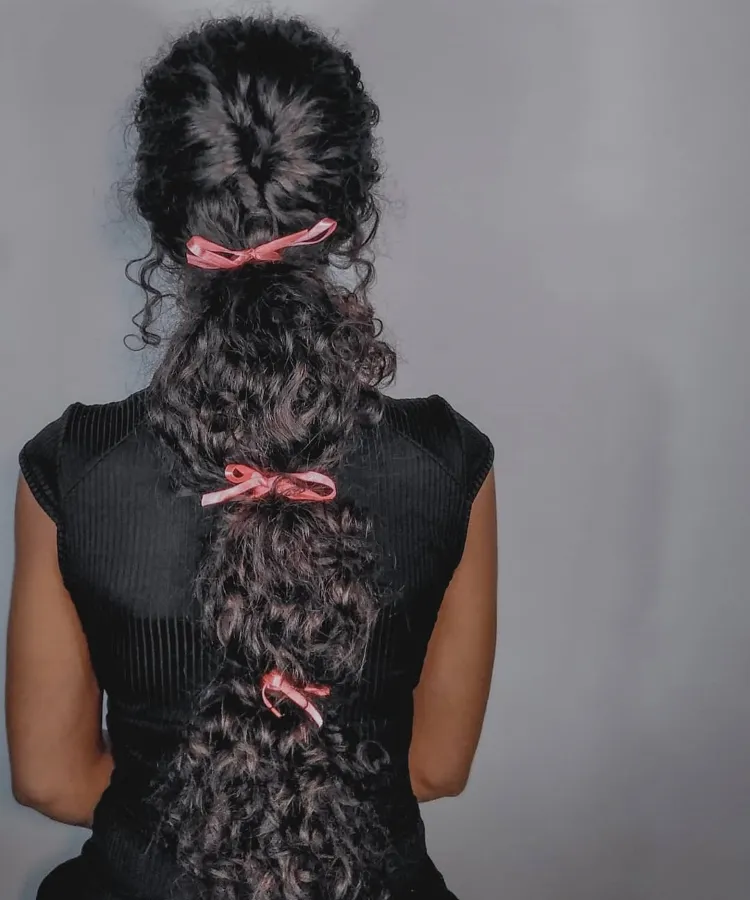 Uma pessoa com cabelos longos, escuros e cacheados é vista de costas, com o cabelo dividido em seções ao longo do comprimento. Cada seção é presa com um pequeno laço de fita rosa. A pessoa veste uma blusa preta de textura canelada. O fundo é um tom uniforme de cinza claro. A iluminação destaca a textura do cabelo e da roupa.



