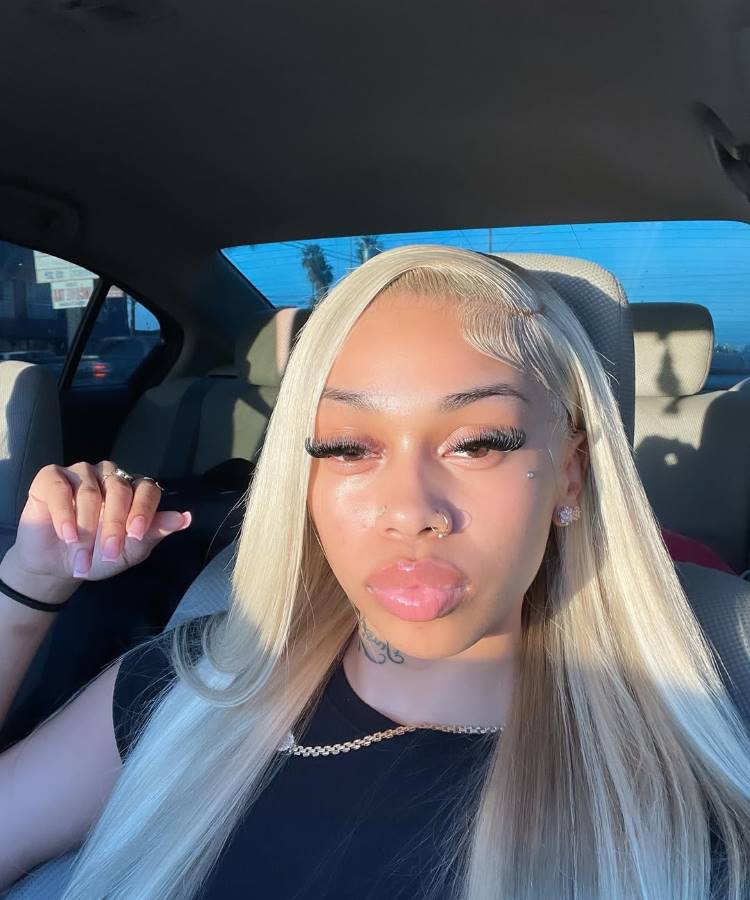 Mulher loira com cabelo liso e baby hairs estilizados. Ela usa maquiagem brilhante, cílios longos, piercing no nariz, colar dourado e camiseta preta. A luz do sol ilumina seu rosto enquanto está sentada dentro de um carro.





