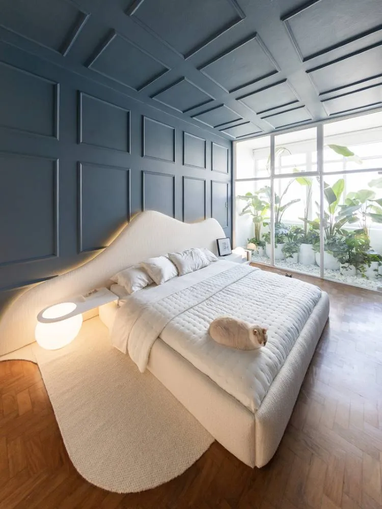 Esta imagem exibe um quarto com uma cama de casal branca e um gato creme deitado sobre ela. A parede atrás da cama é azul-petróleo com painéis. Ao lado da cama há uma luminária branca e uma grande janela revela um jardim interno com plantas verdes.




