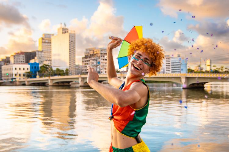 Homem sorridente celebra o carnaval à beira de um rio, vestindo roupas coloridas e segurando uma sombrinha típica. Ao fundo, uma ponte e prédios urbanos sob um céu alaranjado completam o cenário festivo e cultural brasileiro.