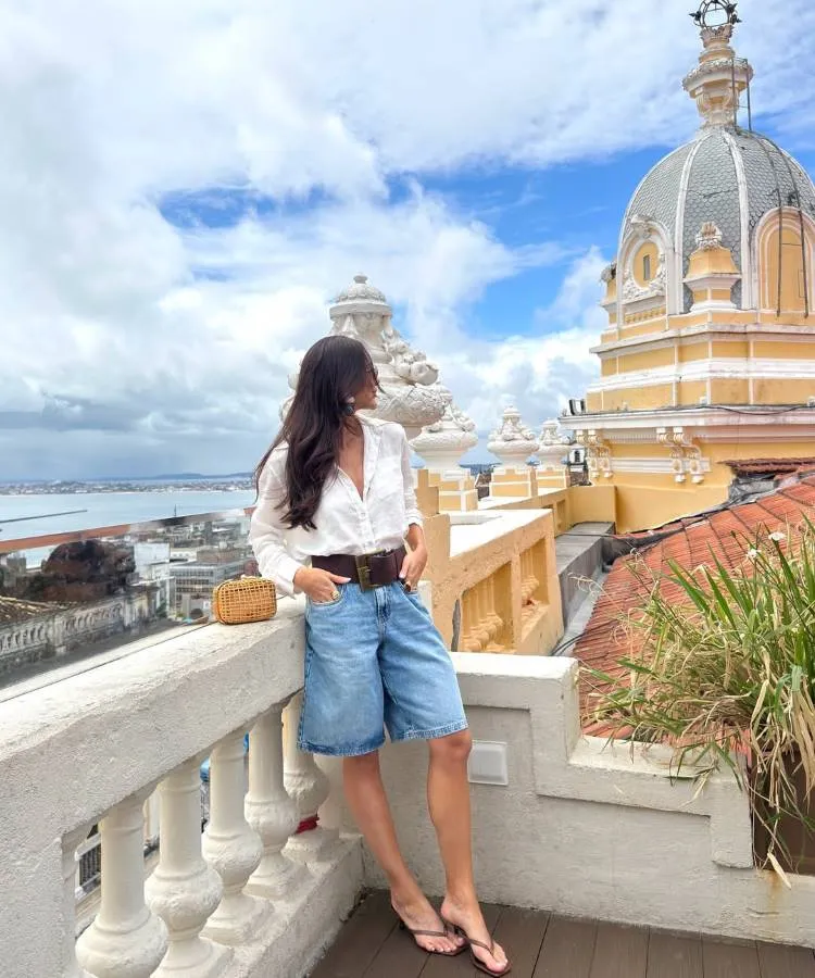 Mulher com cabelos escuros, usando blusa branca, bermuda jeans, cinto largo marrom, bolsa de palha e chinelos. Ela está em uma varanda com vista para o mar e edifícios, de frente para a paisagem.