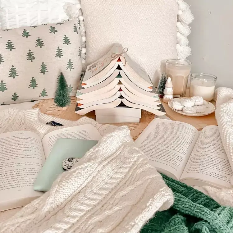Livros empilhados em forma de árvore de Natal, com uma fita no topo. Ao redor, há almofadas com estampas de pinheiros, uma vela acesa, copos de bebida, biscoitos em um prato e um suéter tricotado, criando um ambiente aconchegante e festivo.


