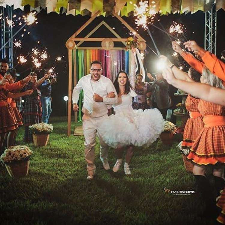 Uma cena animada mostra um casal com trajes de noivos brancos correndo por um espaço festivo ao ar livre, cercado por convidados segurando sparklers. O fundo apresenta um arco colorido e bandeiras, indicando uma alegre celebração de casamento ou uma festa temática de Festa Junina com fogos de artifício.