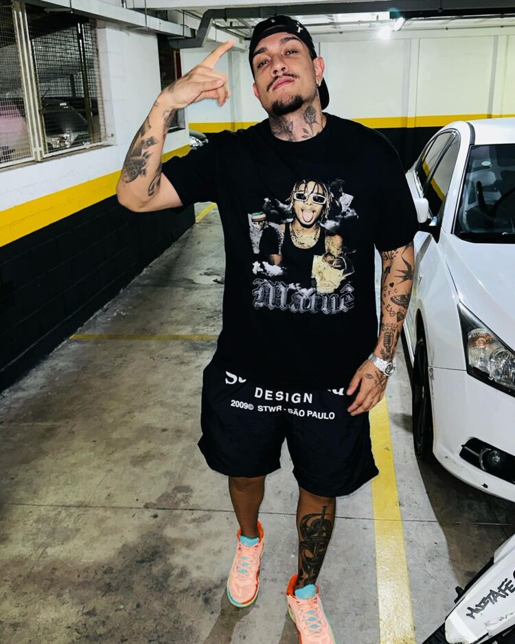 Mc Bin Laden em garagem ao lado de carro branco, usando blusa preta, boné preto para trás, bermuda preta e tênis laranja