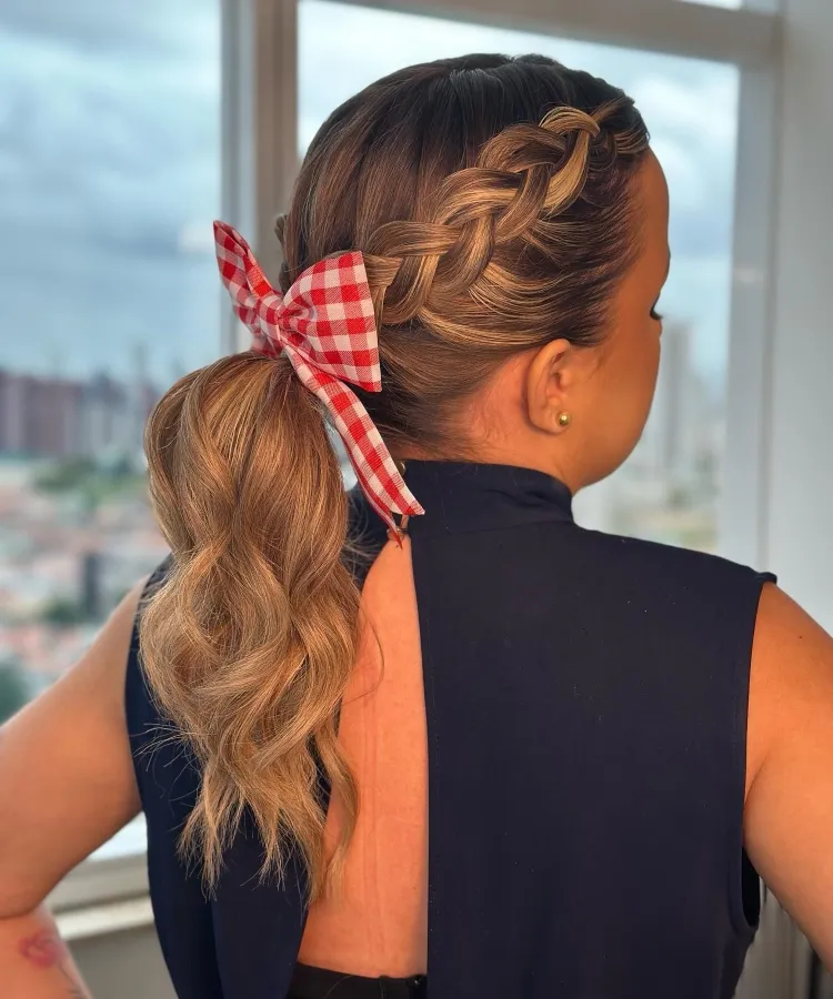 Uma pessoa com cabelos castanhos claros e pele clara é vista de costas, exibindo um penteado elaborado com uma trança lateral que se junta a um rabo de cavalo ondulado. Um grande laço xadrez vermelho e branco adorna a base do rabo de cavalo. A pessoa veste uma blusa preta com uma abertura nas costas. Ao fundo, uma janela com vista para edifícios e o céu sugere um ambiente urbano.

