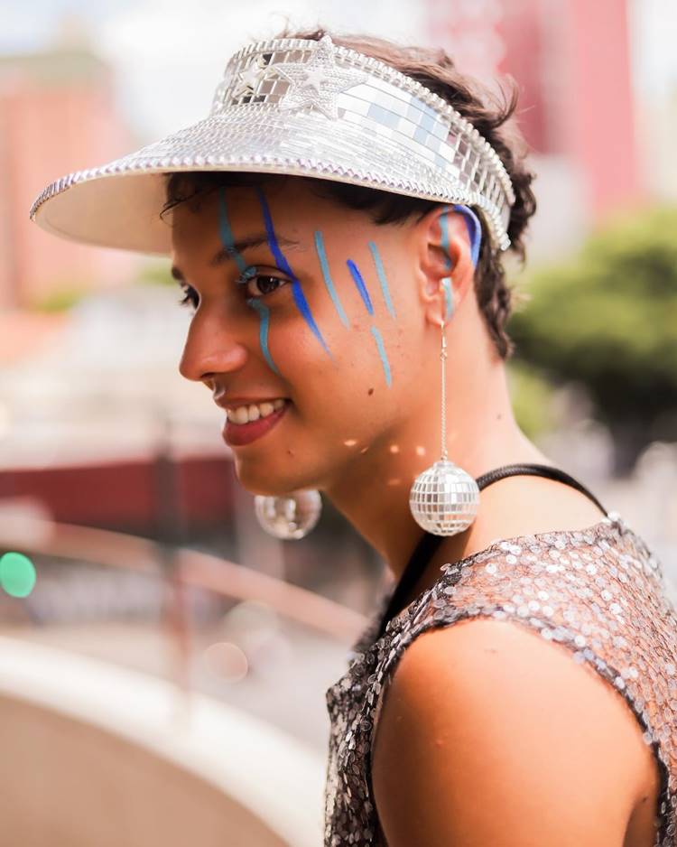 Homem com boné espelhado, regata de miçangas e brincos de globo de festa, com make riscos azuis nos olhos, rosto e orelhas