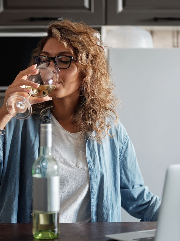 Uma mulher de cabelo encaracolado e óculos, vestindo uma camisa jeans, senta-se à mesa e bebe de uma taça de vinho branco. Uma garrafa de vinho e um laptop estão na mesa.