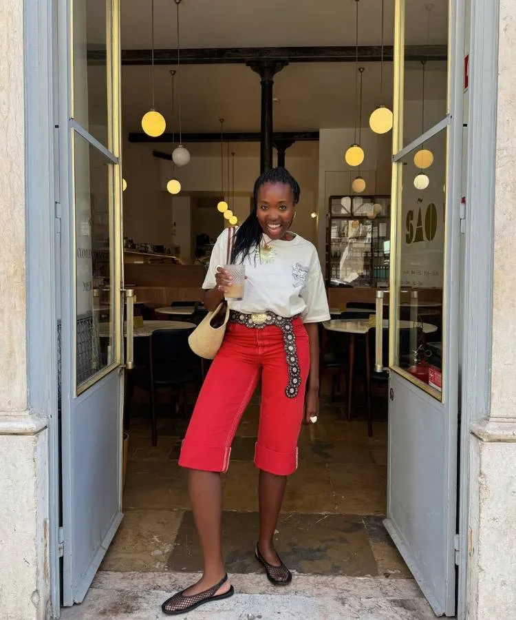 Mulher negra sorridente, com tranças, usando calças capri vermelhas, cinto com detalhes de metal, camiseta creme com estampa floral, bolsa de palha, e sapatilhas de tela pretas. Ela está parada na porta de um restaurante.
