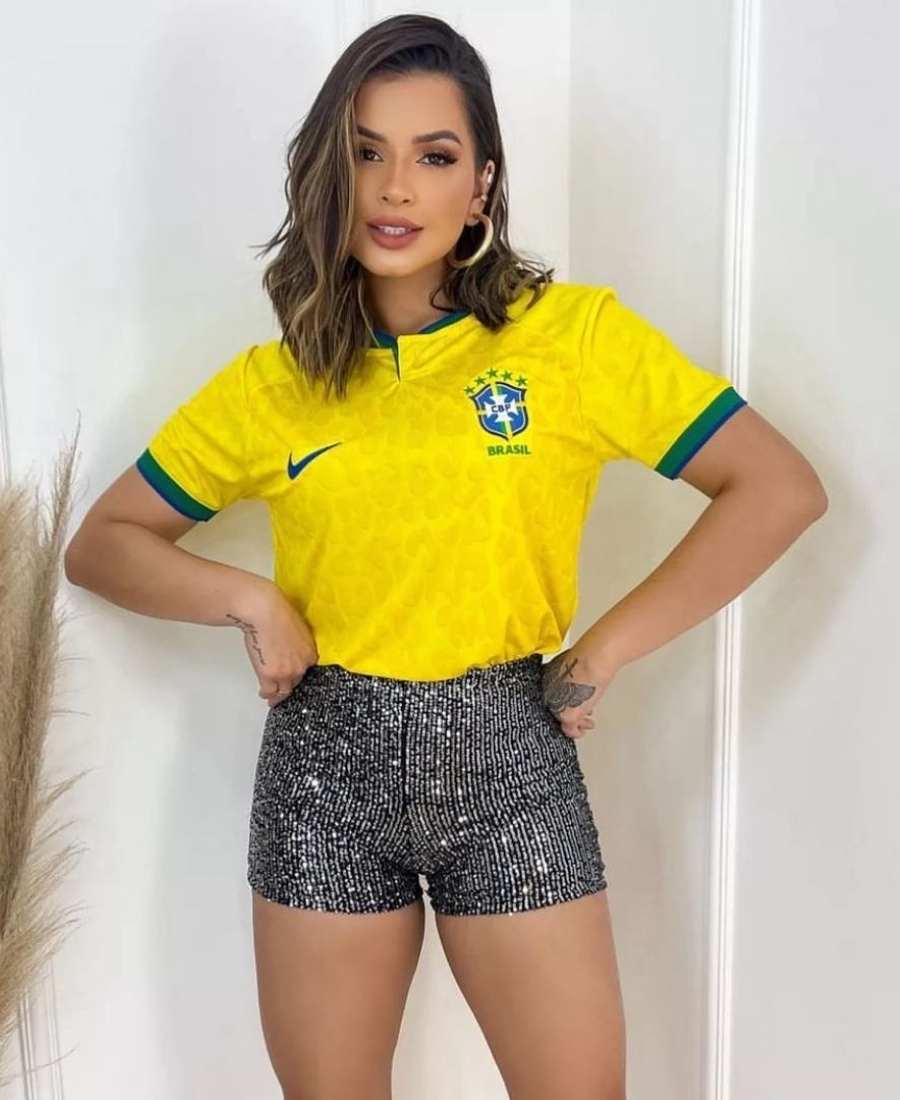 Imagem com fundo de cômodo de casa, com parede e vaso com flor no chão. No centro, mulher usando roupas para a copa do mundo: camiseta amarela do Brasil e short curto preto com lantejoulas prata.