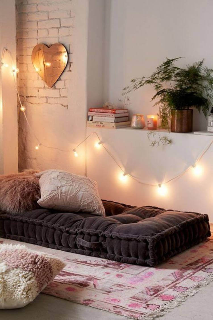 Espaço zen com colchão cinza, almofadas, prateleira com planta, vela e livros, fio com lâmpadas e espelho em formato de coração