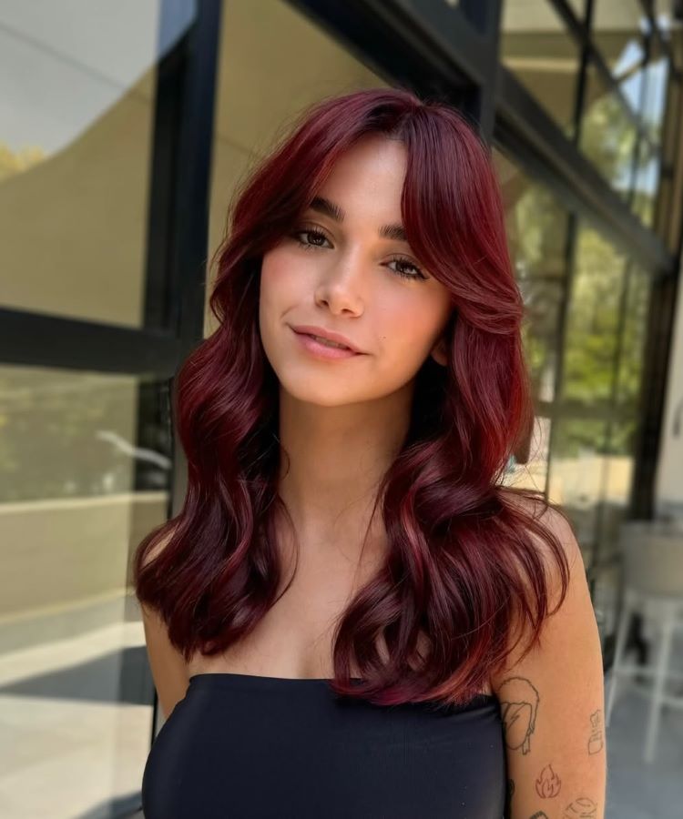 Mulher com cabelos longos, ondulados e brilhosos, destacando cores de cabelo em tom vinho intenso e vibrante. Ela veste uma blusa preta tomara-que-caia, exibe um sorriso suave e está em um ambiente externo iluminado, com vidro ao fundo.