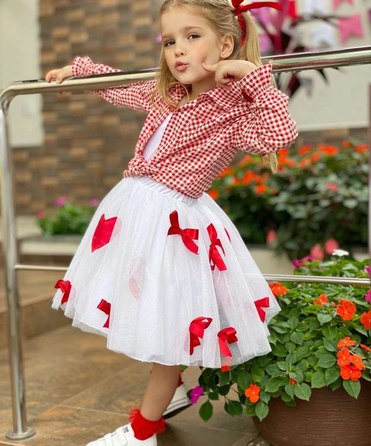 Menina usando camisa xadrez vermelha com saia branca e laços vermelhos para festa junina.
