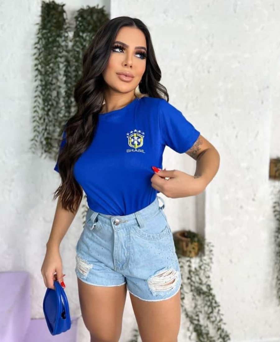 Imagem com fundo parede branca com fvaso de plantas. No centro, mulher usando roupas Brazilcore: camiseta azul do Brasil + short jeans + bolsa azul.
