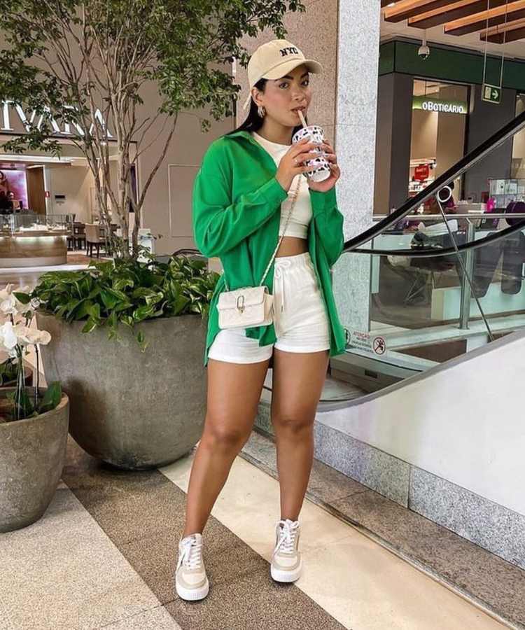 Imagem com fundo de escada de shopping, lojas e plantas. No centro, mulher usando cropped branco, short, branco, bolsa branca, tênis branco, boné bege e blazer verde